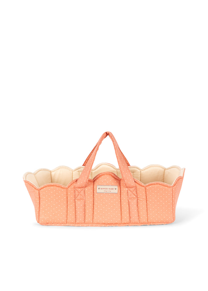 The Konges Sloejd Doll Lift - Rose Dot is a peach-hued quilted cotton baby basket featuring white polka dots, scalloped edges, two convenient handles, and a small label on the front. OEKO-TEX certified for safety and comfort, this delightful doll carrier is shown empty against a white background.