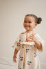 A young child smiles, sipping from a Konges Sloejd Glitter Cup - Cherry against a light background. She wears a cream dress with red cherries, her curly hair in a ponytail. These fun glitter cups make every drink special.
