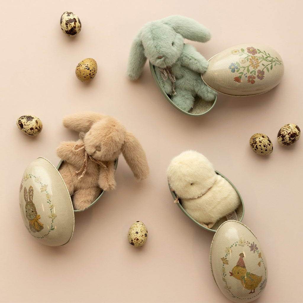 A soft pastel background with two plush rabbits, a plush chick, and several decorative eggs, including a Maileg Easter Egg, arranged in a gentle, festive Easter theme.