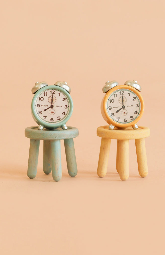 Two small alarm clocks, one blue and one yellow, are displayed atop the Maileg Stool Set, Mouse - Mint/Light Yellow, their beech wood construction and vintage paint finish adding charm against a beige background.