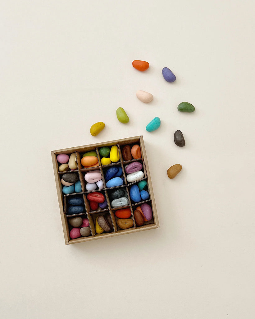 A small, square box containing neatly arranged, colorful Easter Basket Sets in separate compartments, with a few stones scattered outside the box on a light background.
