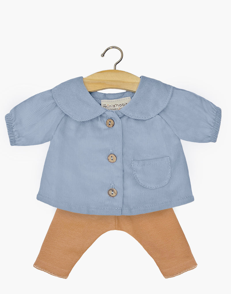 The Minikane Doll Clothing set includes a light blue blouse with long sleeves, adorned with three brown buttons and a small front pocket. The hand washable blouse is displayed on a wooden hanger and comes paired with light brown Sugar Jersey leggings.