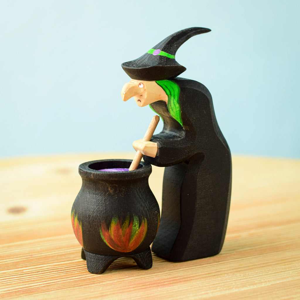 The Bumbu Handmade Wooden Brewing Witch and Cauldron Set features a small wooden witch with a green streak in her hair and a pointy hat, stirring a black cauldron with painted flames—a charming Montessori Waldorf toy.