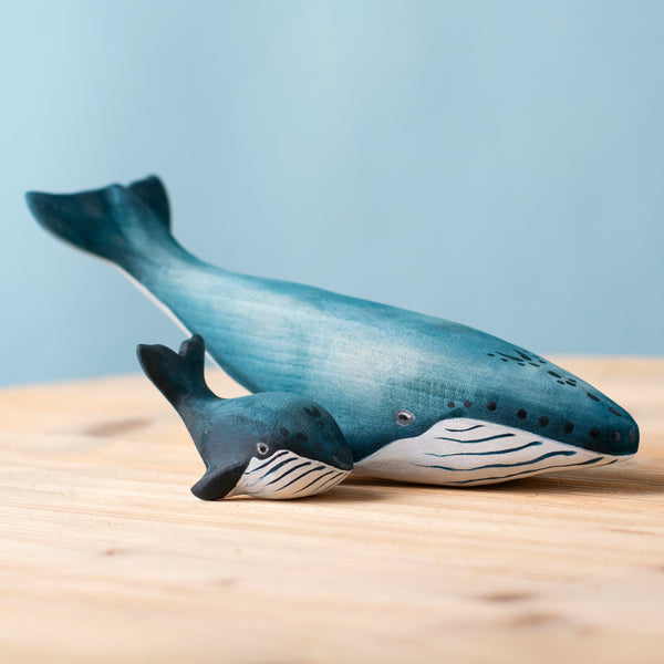 The Bumbu Handmade Wooden Blue Whale Family Set features two painted wooden whale figurines, large and small, on a light wood surface against a pale blue background—sustainable toys ideal for Montessori and imaginative play.