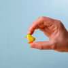 A hand holds the Bumbu Handmade Wooden Chick, a small yellow wooden figurine, set against a light blue background.