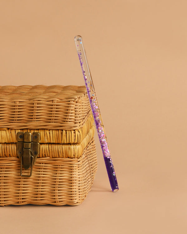 The Konges Sloejd Magical Glitter Wand - Unicorn, filled with purple sparkles, rests at an angle against a closed wicker basket with a metal latch, set on a beige background.