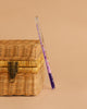 The Konges Sloejd Magical Glitter Wand - Unicorn, filled with purple sparkles, rests at an angle against a closed wicker basket with a metal latch, set on a beige background.