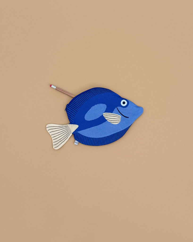 A Surgeon Fish Pencil Case, crafted from blue cotton with a white tail and fins, rests on a beige surface. A pencil peeks out from its zippered opening.