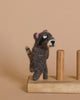 The Raccoon Finger Puppet, crafted entirely from 100% wool in brown and black tones, is a delightful creation perched on a wooden stand with two pegs, set against a soft beige background.