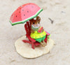 A Wee Forest Folk Tiny Collectible features a hand-sculpted mouse in a yellow dress on a red chair in the sand, reading under a watermelon umbrella with a watermelon slice—Perfect Beach Day!.