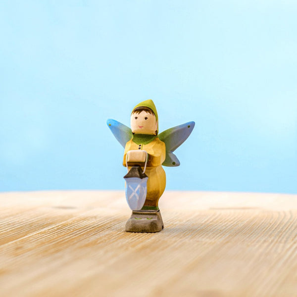 The Bumbu Handmade Wooden Winged Elf with Lamp Set, featuring a small elf in a yellow outfit with blue wings and a lantern, stands on a light wooden surface against a soft blue background.