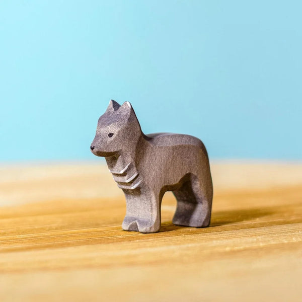 The Bumbu Handmade Wooden Wolf Cub, a small handcrafted wooden toy shaped like a gray wolf, stands on a light wooden surface with a soft blue background.
