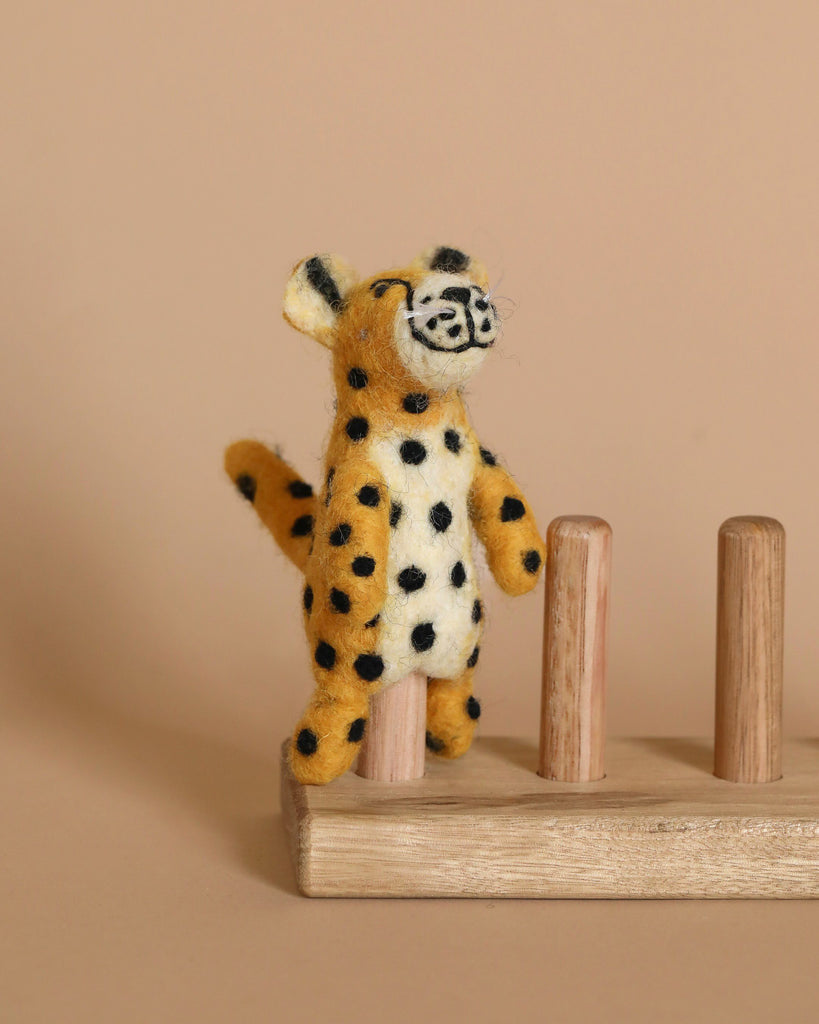 A Cheetah Finger Puppet with black spots stands on a wooden pegboard toy against a beige background. The cheetah, featuring a smiling face and crafted from needle felt, is positioned upright on one of the pegs.