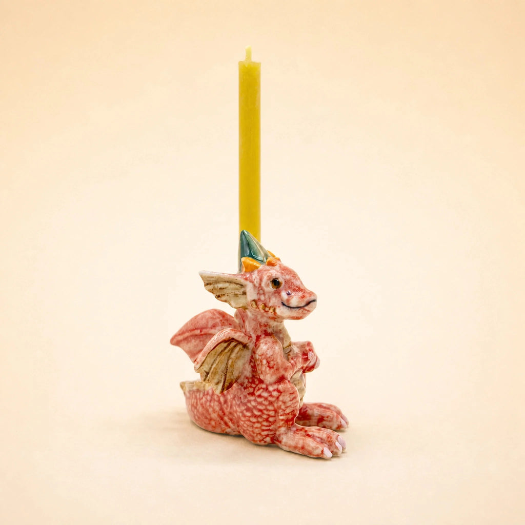 A yellow candle is displayed in the Dragon Cake Topper—a handcrafted collectible holder shaped like a small red dragon with green horns, gold wings, and a textured body—set against a plain beige background.