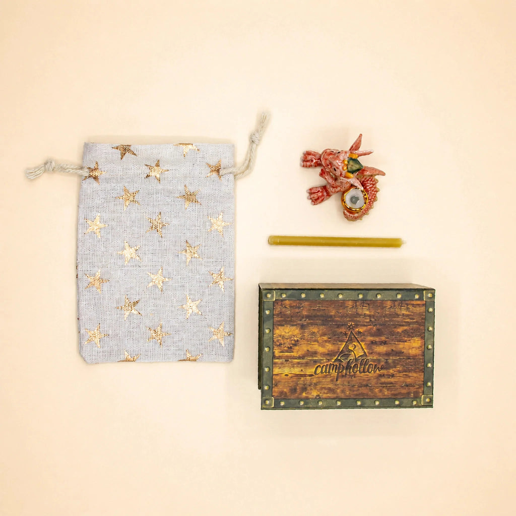 A small gray star-patterned pouch, a yellow pencil, a wooden keepsake treasure chest with “Camp Hollow” on the lid, and a red Dragon Cake Topper - Final Sale are arranged on a cream background.