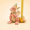Introducing the Dragon Cake Topper - Final Sale: a small, whimsical pink dragon figurine sporting wings and a party hat, playfully holding a yellow beeswax birthday candle in a cracked eggshell with its tail—making it the perfect handcrafted collectible for your celebration.
