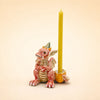 A small, red, ceramic Dragon Cake Topper with a yellow crown sits beside a yellow candle held in a cracked egg-shaped holder. This handcrafted collectible, offered as a final sale item, would make an enchanting addition to any keepsake treasure chest. The background is light beige.