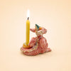 A whimsical ceramic holder shaped like a pink dragon with wings and small horns—reminiscent of a handcrafted collectible—displays the Dragon Cake Topper (Final Sale) against a plain beige background. A yellow candle burns inside, highlighting its playful design.
