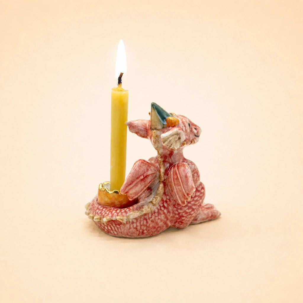 A whimsical ceramic holder shaped like a pink dragon with wings and small horns—reminiscent of a handcrafted collectible—displays the Dragon Cake Topper (Final Sale) against a plain beige background. A yellow candle burns inside, highlighting its playful design.