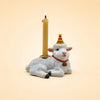 A small porcelain goat figurine wearing a red and yellow party hat holds a lit candle on its back. Perfect as the Goat Cake Topper – Final Sale, this handcrafted birthday decoration stands out against the plain, light beige background.