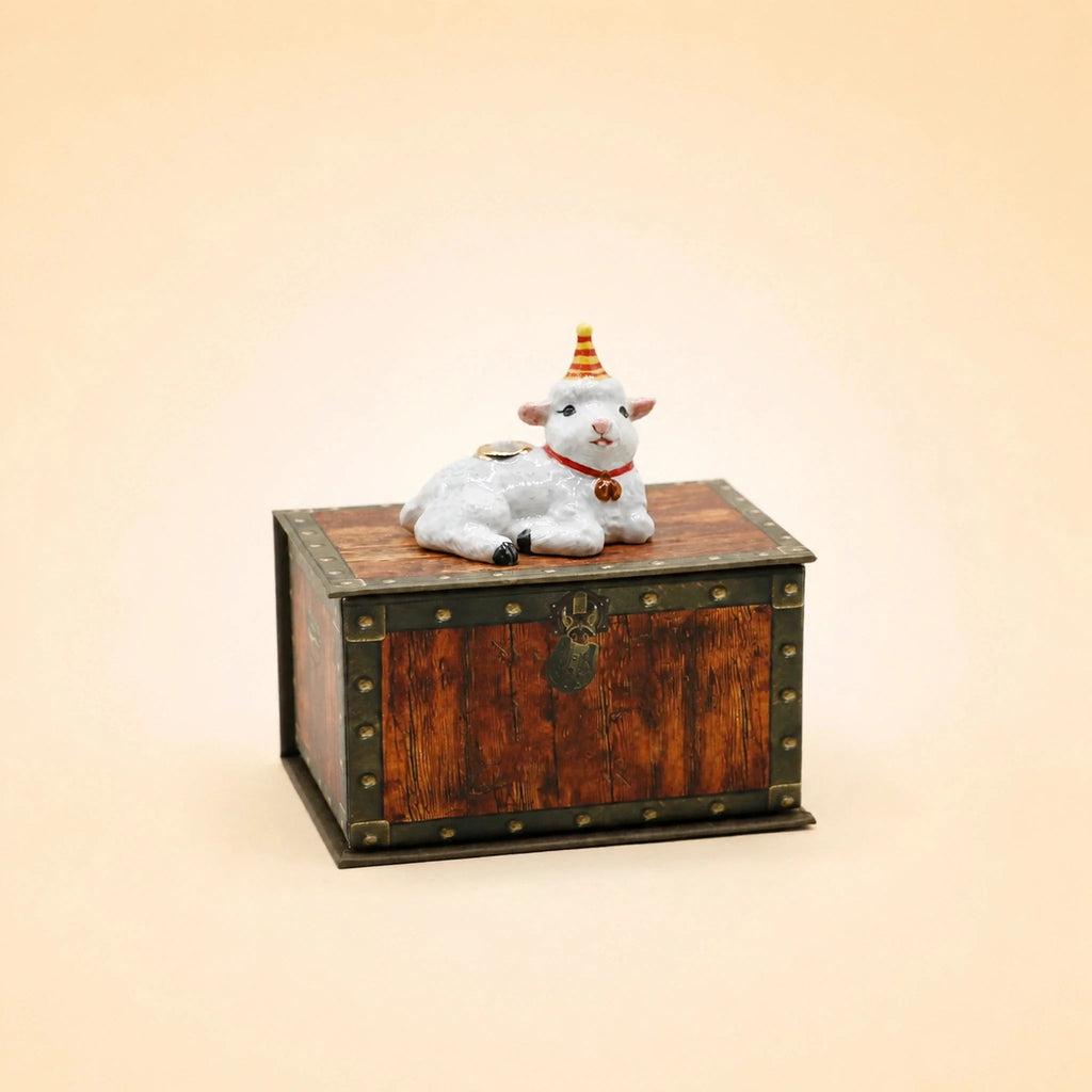 A small wooden chest with metal accents sits on a light background, topped with the Goat Cake Topper collectible keepsake figurine.