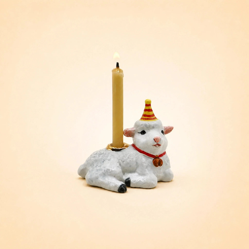 A small porcelain goat figurine, perfect as a cake topper, wears a red and yellow party hat and has a red collar with a bell. The goat holds a lit beige candle on its back and is photographed against a plain, light background. Product Name: Goat Cake Topper - Final Sale.