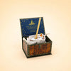A small wooden chest with an open lid reveals a collectible keepsake—a Goat Cake Topper (Final Sale), carefully wrapped and accompanied by a stick. The inside cover is dark blue adorned with gold stars and text, while the background is light beige.