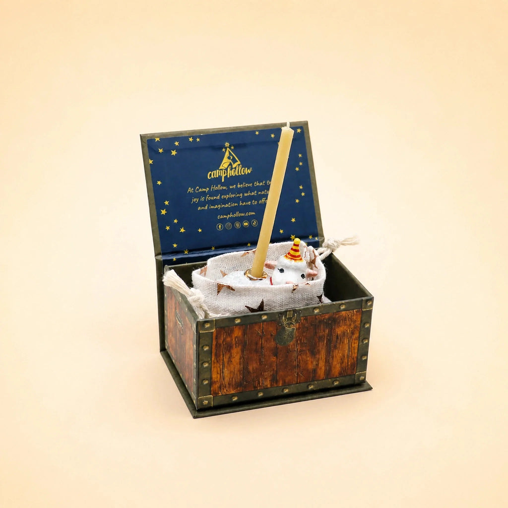 A small wooden chest with an open lid reveals a collectible keepsake—a Goat Cake Topper (Final Sale), carefully wrapped and accompanied by a stick. The inside cover is dark blue adorned with gold stars and text, while the background is light beige.