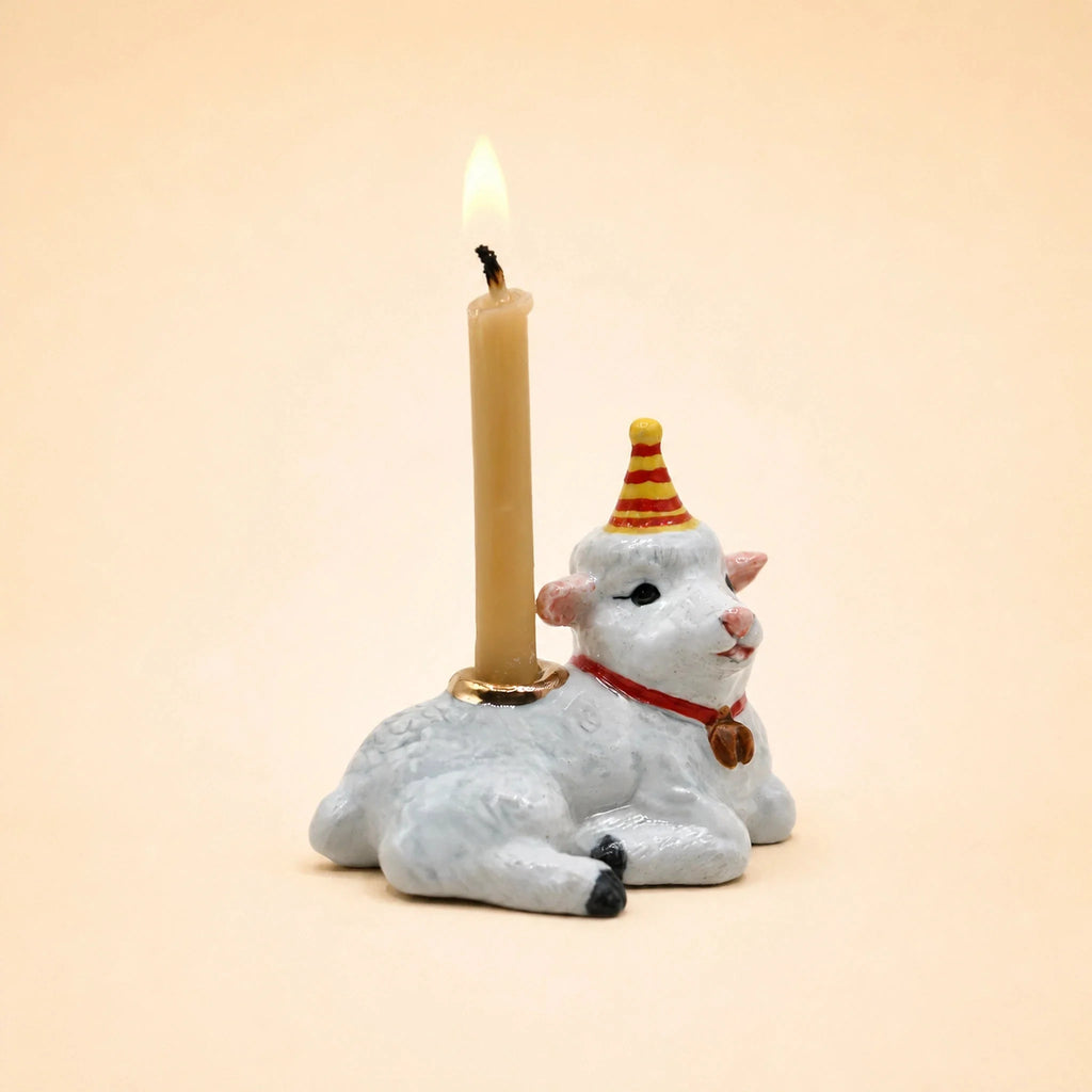 A handcrafted Goat Cake Topper, featuring a white goat figurine sporting a red and yellow party hat and carrying a lit beige candle on its back, makes an adorable birthday decoration against a plain light background.