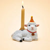 The Goat Cake Topper - Final Sale features a charming white goat figurine adorned with a red and yellow party hat, holding a lit beige candle on its back. Accentuated with a red collar and small bell, this handcrafted birthday decoration stands out against a soft light peach background, making it a delightful collectible keepsake.