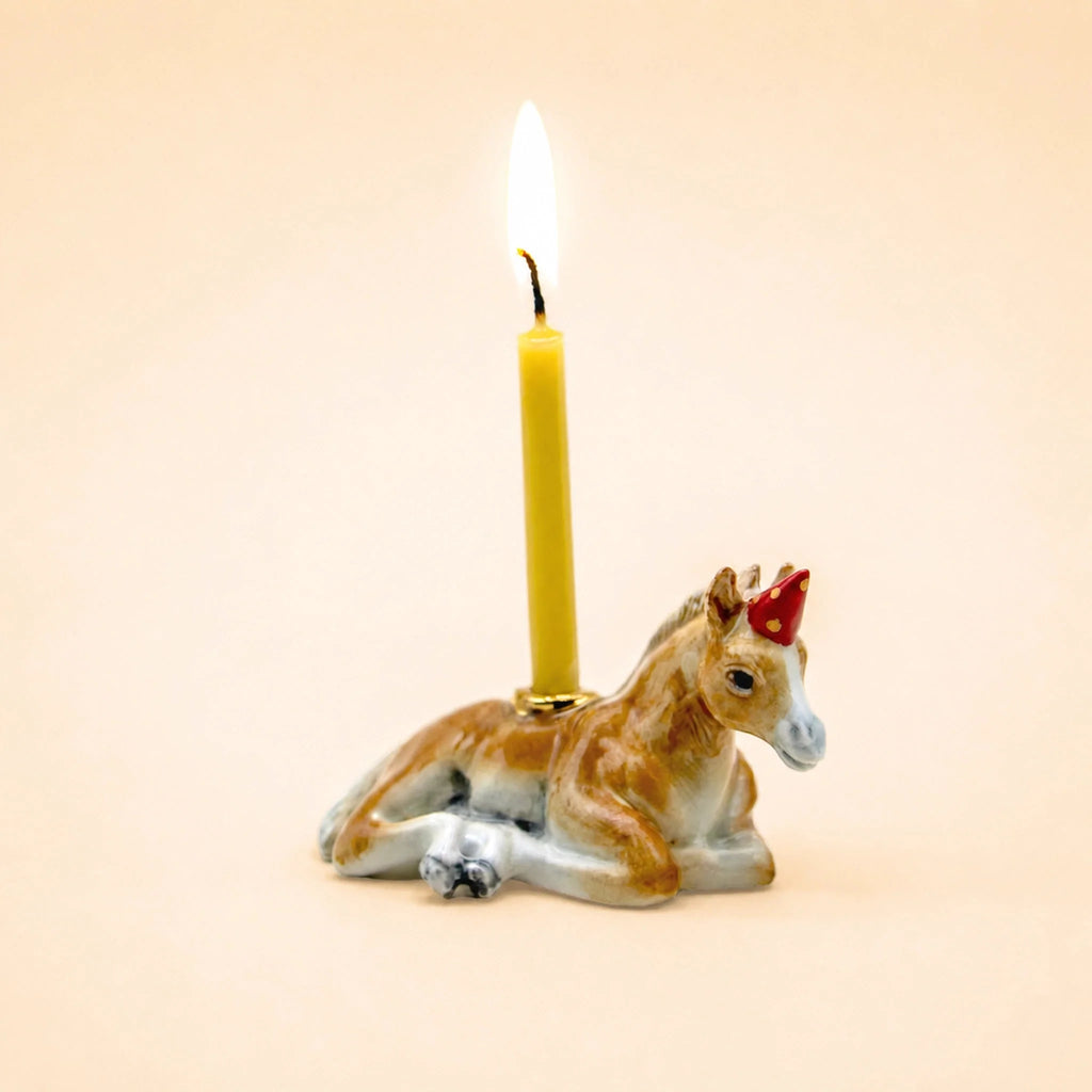 A yellow candle is placed on the Horse Cake Topper—a ceramic horse figurine featuring subtle gold accents and a small red party hat—set against a plain beige background.