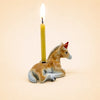 Here’s a rewritten sentence using your product data: The “Horse Cake Topper - Final Sale” is a small porcelain figurine depicting a brown donkey wearing a red party hat and lying down, with a lit yellow candle inserted vertically on its back, set against a plain beige background.