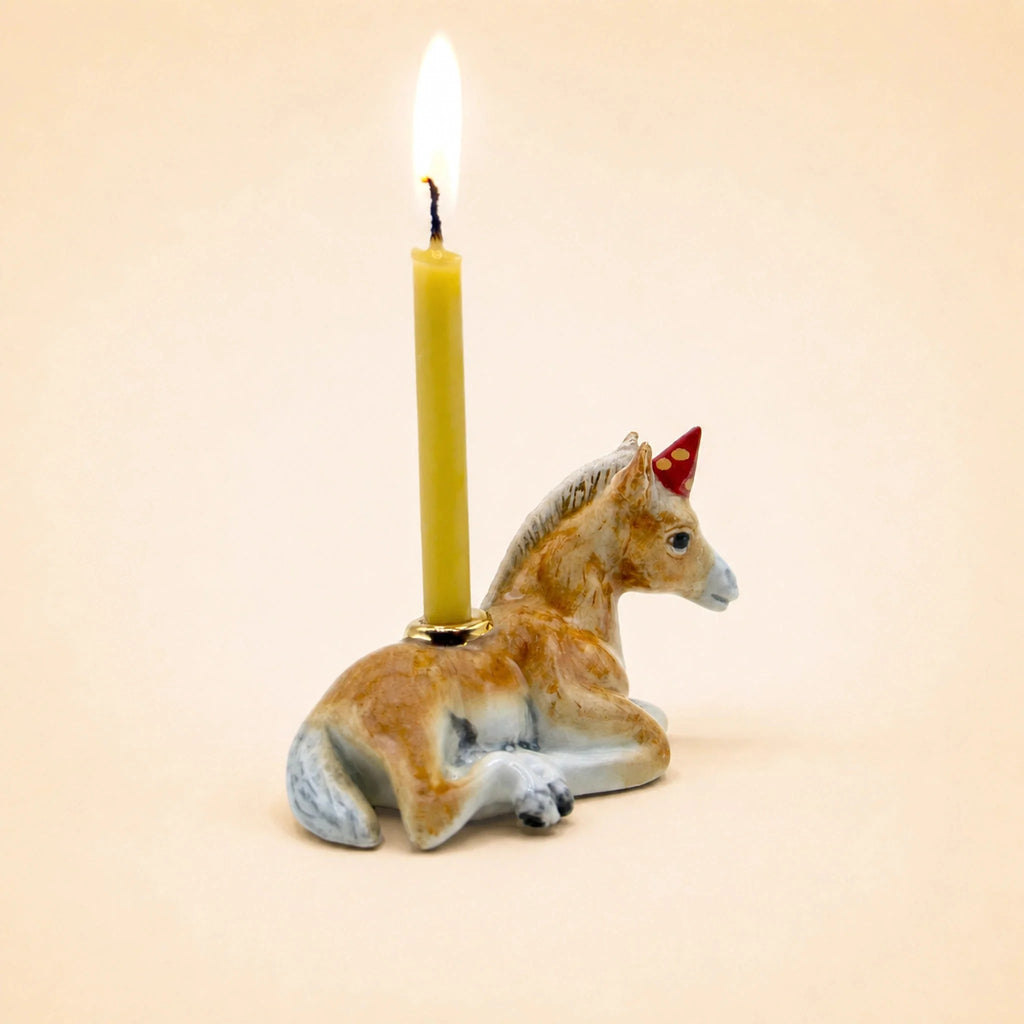 Here’s a rewritten sentence using your product data: The “Horse Cake Topper - Final Sale” is a small porcelain figurine depicting a brown donkey wearing a red party hat and lying down, with a lit yellow candle inserted vertically on its back, set against a plain beige background.