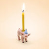 A small handcrafted Pig Cake Topper - Final Sale features a collectible ceramic pig wearing a blue and white striped party hat and holding an upright, lit yellow birthday candle on its back, set against a plain light beige background.