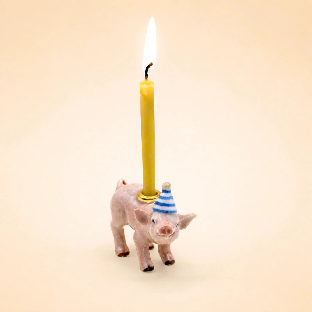 A small handcrafted Pig Cake Topper - Final Sale features a collectible ceramic pig wearing a blue and white striped party hat and holding an upright, lit yellow birthday candle on its back, set against a plain light beige background.