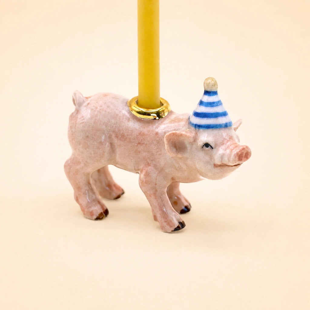 Introducing the Pig Cake Topper - Final Sale: a petite ceramic pig figurine, handcrafted with care, featuring a blue and white striped party hat and carrying a yellow candle on its back accented by a gold ring. This delightful porcelain collectible is perfect as an adorable cake topper or a charming keepsake birthday decoration.