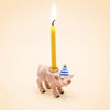 The Pig Cake Topper - Final Sale features a small pig figurine wearing a blue-and-white striped party hat and holding a lit yellow candle on its back, making it an ideal keepsake birthday decoration against a plain beige background.