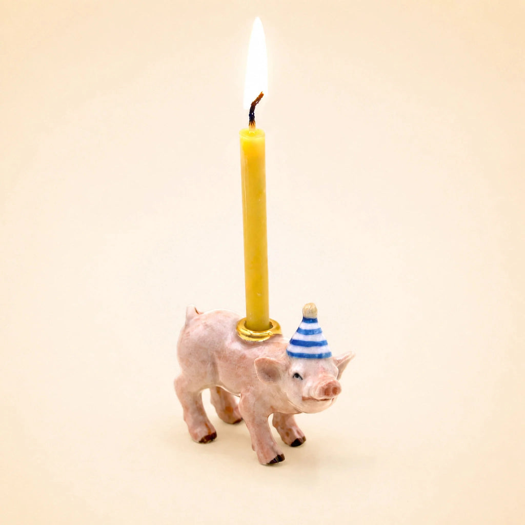 The Pig Cake Topper - Final Sale features a small pig figurine wearing a blue-and-white striped party hat and holding a lit yellow candle on its back, making it an ideal keepsake birthday decoration against a plain beige background.