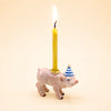 A small handcrafted Pig Cake Topper figurine wears a blue-and-white party hat and carries a lit yellow candle on its back, set against a light beige background.