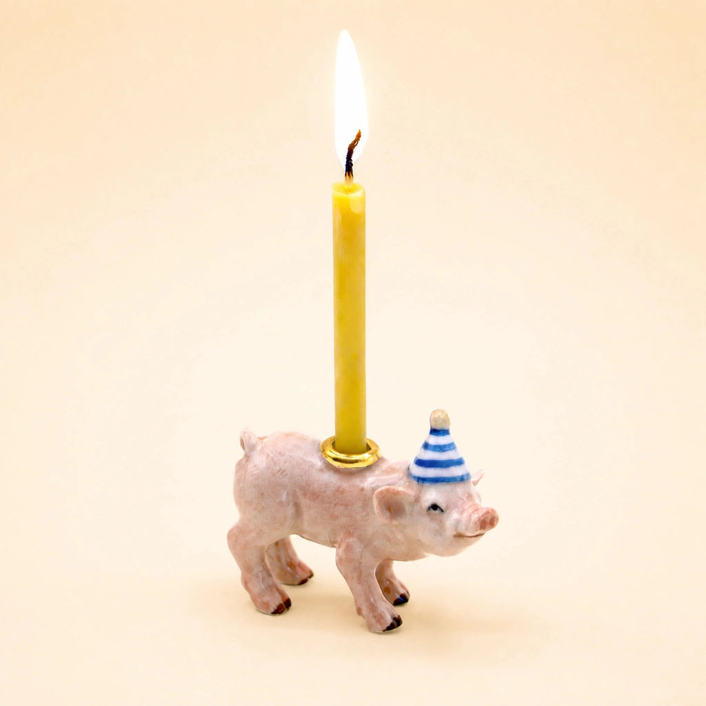 A small handcrafted Pig Cake Topper figurine wears a blue-and-white party hat and carries a lit yellow candle on its back, set against a light beige background.