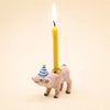 The Pig Cake Topper - Final Sale features a small pig figurine adorned with a blue and white striped party hat, holding a lit yellow candle on its back against a plain light background—making it an adorable keepsake birthday decoration or porcelain cake topper.