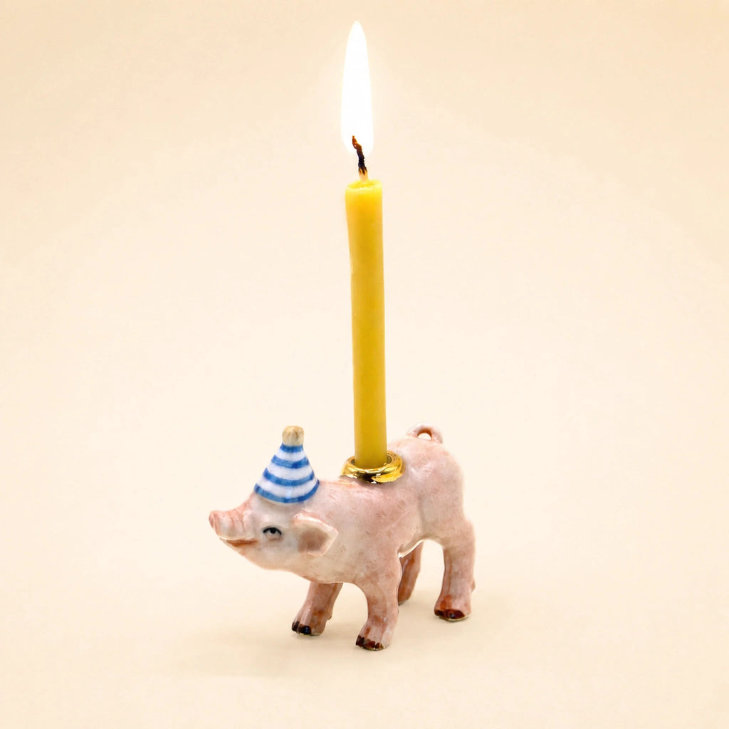 The Pig Cake Topper - Final Sale features a small pig figurine adorned with a blue and white striped party hat, holding a lit yellow candle on its back against a plain light background—making it an adorable keepsake birthday decoration or porcelain cake topper.