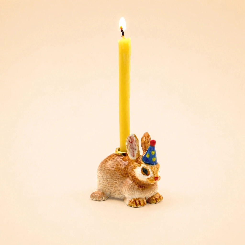 The Bunny Rabbit Cake Topper - Final Sale features a ceramic bunny figurine holding a lit yellow candle and wearing a blue party hat with yellow dots, set against a plain beige background.