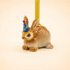 Bunny Rabbit Cake Topper - Final Sale: This handcrafted ceramic bunny figurine features a blue party hat adorned with yellow dots and a red tip, and is holding a yellow candle on its back. Displayed against a plain light background, it makes a charming collectible or unique cake topper.