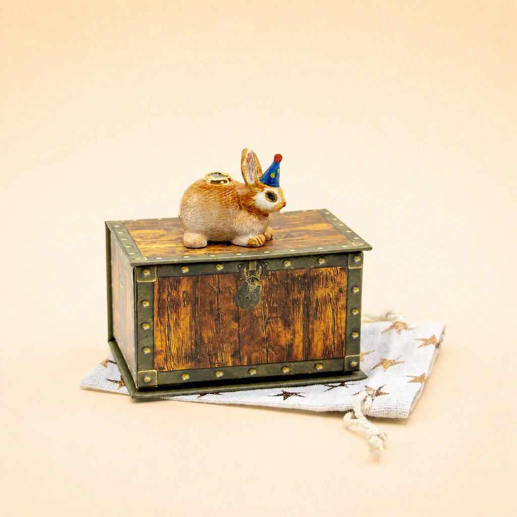 A small handcrafted collectible wooden chest sits on a star-patterned cloth. On the chest rests the Bunny Rabbit Cake Topper, featuring a figurine of a rabbit wearing a blue party hat.