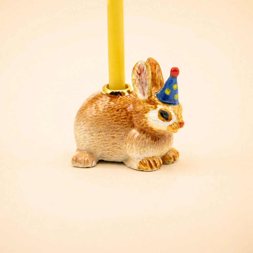 The Bunny Rabbit Cake Topper - Final Sale features a handcrafted collectible ceramic bunny figurine wearing a blue party hat with yellow dots and carrying a yellow candle on its back, displayed against a plain, light background.