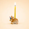 A handcrafted Bunny Rabbit Cake Topper figurine, featuring a ceramic rabbit in a blue party hat holding a lit yellow candle on its back, is set against a plain light background.