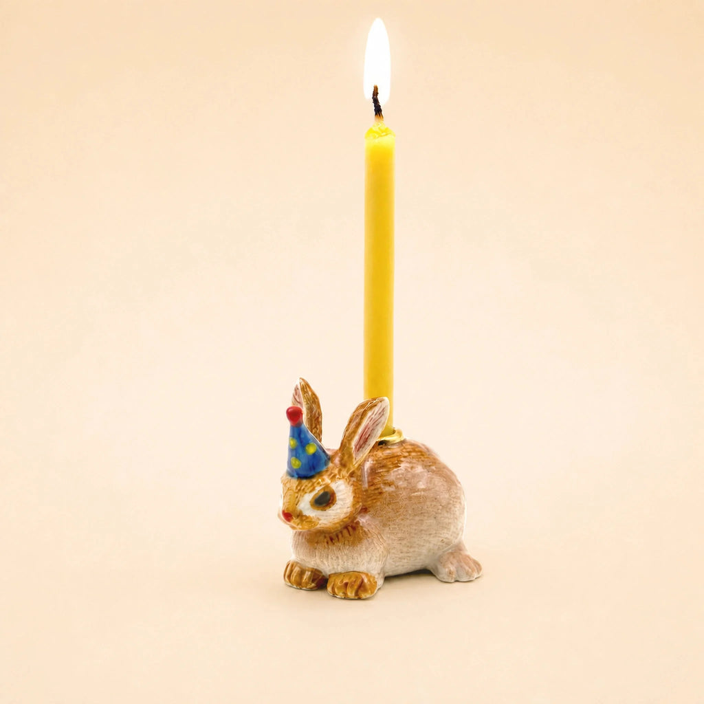 The Bunny Rabbit Cake Topper – Final Sale is a handcrafted collectible ceramic rabbit wearing a blue party hat and holding a lit yellow candle on its back, displayed against a plain beige background.