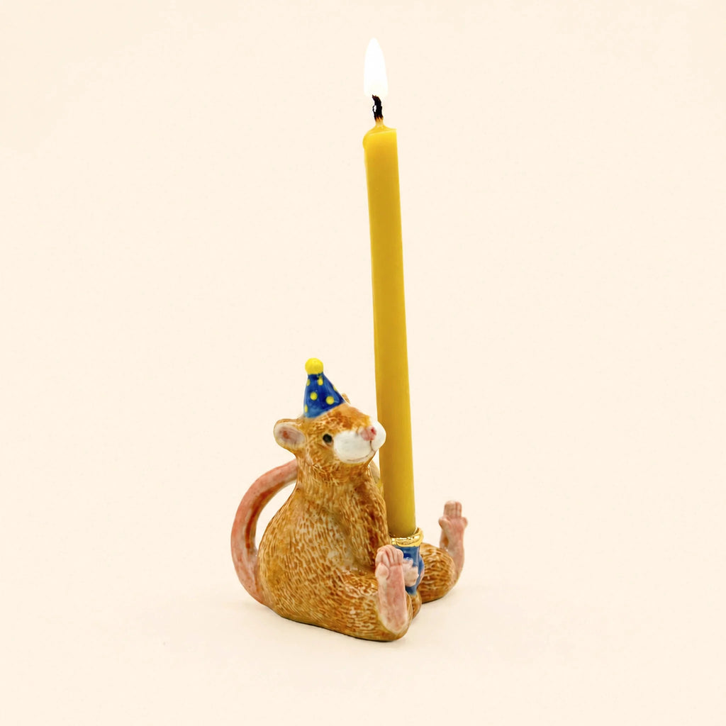A hand-painted birthday decoration, the Mouse Cake Topper - Final Sale is a ceramic figurine featuring a mouse wearing a blue party hat and holding an upright lit yellow candle while sitting on its back feet against a plain light background.