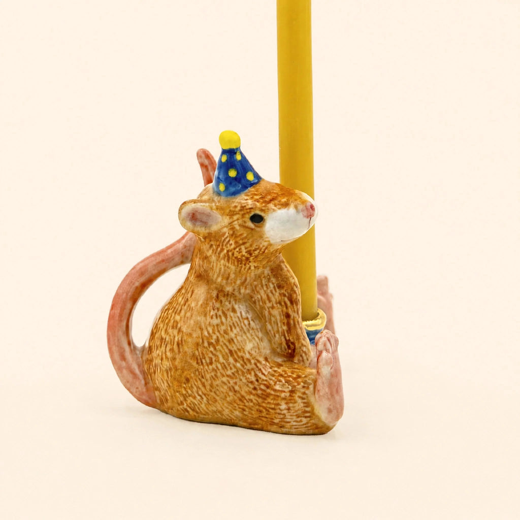 Certainly! Here’s a rewritten sentence using the product data: The Mouse Cake Topper – Final Sale is a hand-painted ceramic figurine featuring a mouse wearing a blue party hat with yellow dots, holding a yellow candle upright with its feet and tail, set against a plain light background.